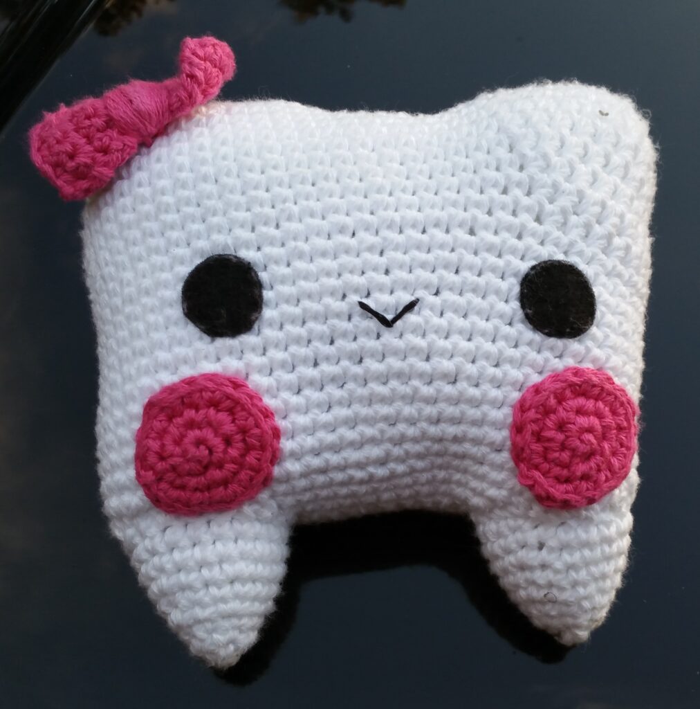 crocheted amigurumi tooth fairy pouch with pink bow at top left, with round black eyes, small thread mouth and two rosy pink cheeks